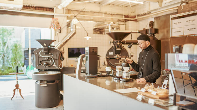 A café worker in black attire and a hat pouring coffee amidst a white-tiled space filled with industrial machinery.