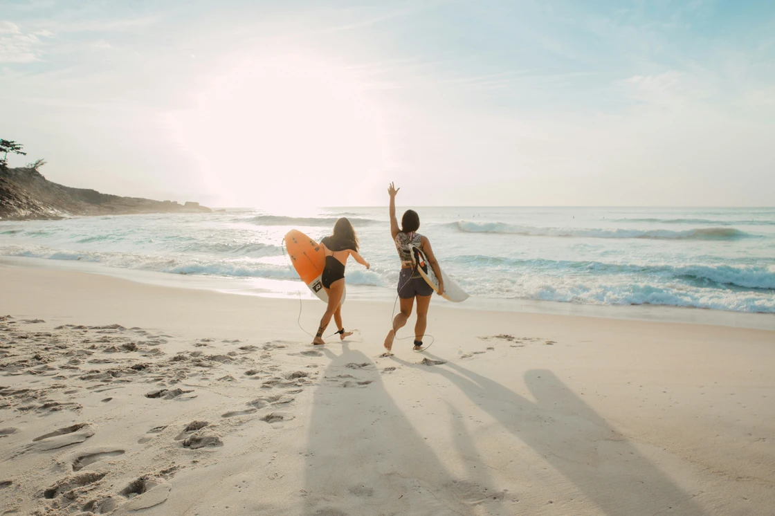 two woman at the beach holding a surfboard under their arm walking to the water